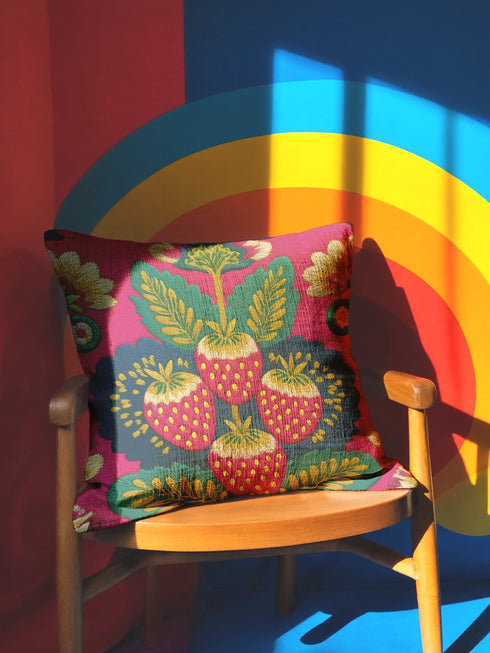 A square throw pillow with a vibrant, folk art-inspired design. The pillow's background is a bright, saturated fuchsia pink. The central design features four stylized strawberries arranged in a cluster, rendered in a way that mimics thick, textural embroidery. Each strawberry is red. The overall aesthetic is colorful, eclectic, and maximalist, with a distinct retro or 70s feel. The pillow cover has a visible linen-like texture.