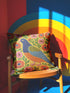 Square faux embroidery throw pillow cover with a large stylized folk art bird facing left. The bird has a cobalt blue head, orange belly, light blue tail, and pink legs, set on a bright mustard yellow background scattered with pink and red daisy flowers on trailing green vines. The smooth, linen-feel polyester uses a printed trompe loeil effect to mimic raised thread needlework. Bold maximalist folk art bird pillowcase and cottagecore cushion cover for eclectic, colorful, dopamine decor homes.