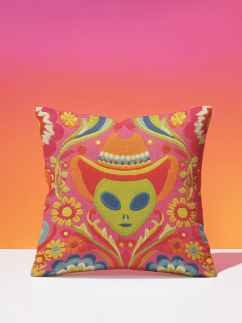A vibrant pink square throw pillow featuring a green alien head wearing an orange cowboy hat. The alien is surrounded by colorful retro floral patterns in yellow, blue, and red. The style combines space cowgirl aesthetic with maximalist dopamine decor. The fabric has a premium linen texture. The cushion sits on a bright blue sofa, showcasing its bold, funky design. Perfect for weirdcore, sci-fi, or western themed rooms.