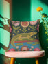 A vibrant square throw pillow cover featuring a green alligator illustration on a dark charcoal background. The design mimics the texture of vintage needlepoint embroidery on faux linen fabric. The alligator is accented with lime green scales and hot pink floral and lip motifs, creating a maximalist junglecore aesthetic. The pillow is shown on a furniture piece, highlighting the vivid cherry red and neon accents typical of dopamine decor and swampcore styles.