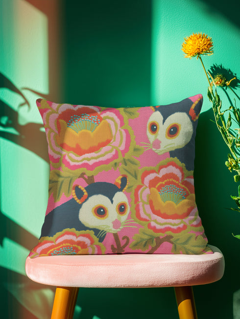 A square linen-feel throw pillow with a vibrant, maximalist print on a bright pink background. The pattern features two large, illustrated possums (or sugar gliders). The animals are nestled amongst large, 70s-style retro floral designs in shades of orange, marigold yellow, and turquoise, with avocado green leaves. The pillow has a textured, woven appearance, mimicking a high-end linen fabric. The double-sided print is colorful, eclectic, and groovy, with a distinct vintage 70s folk art feel.