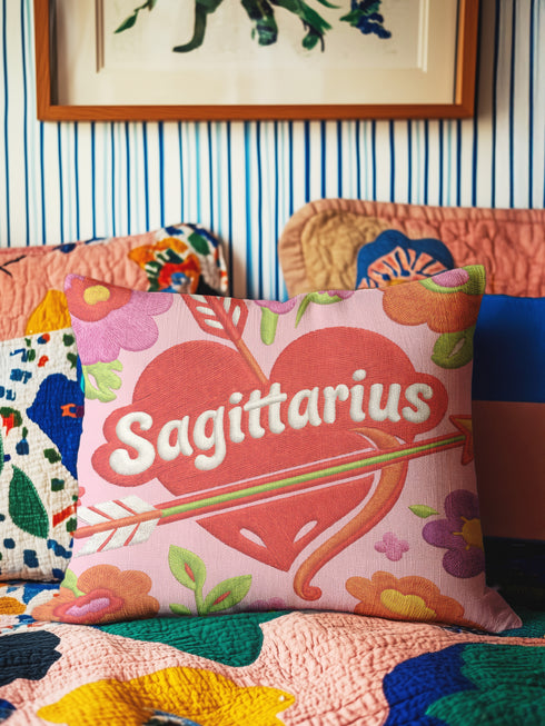 A square decorative throw pillow featuring a light pink background with a trompe-l'œil faux-embroidery print. A large red heart sits in the center, pierced by a green and white arrow. The word Sagittarius is printed across the heart in a puffy white retro font with a red drop shadow. The central design is surrounded by scattered vintage-style flowers in vibrant shades of purple, orange, and yellow with bright green leaves. The texture is a flat, high-fidelity printed illusion.