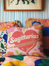 A square decorative throw pillow featuring a light pink background with a trompe-l'œil faux-embroidery print. A large red heart sits in the center, pierced by a green and white arrow. The word Sagittarius is printed across the heart in a puffy white retro font with a red drop shadow. The central design is surrounded by scattered vintage-style flowers in vibrant shades of purple, orange, and yellow with bright green leaves. The texture is a flat, high-fidelity printed illusion.