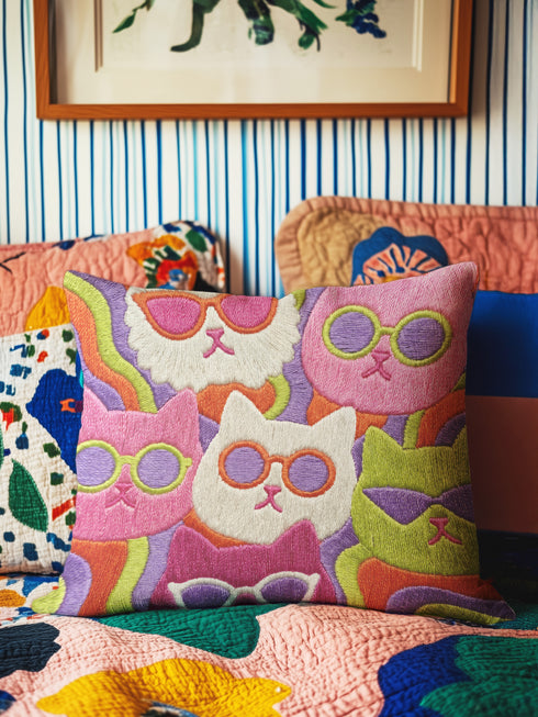 Square throw pillow cover packed with a dense repeating pattern of cat faces in hot pink, lime green, purple, and white, every cat wearing oversized retro sunglasses. Wavy psychedelic bands of orange, yellow, and green weave through the background. The flat 2D print mimics heavy textured crewel needlepoint on a smooth, linen-feel fabric. Reversible same design front and back with a hidden zipper. Funky maximalist cat decor and quirky apartment warming gift, available in 18 by 18 inch and 22 by 22 inch, cove