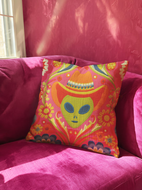 A vibrant pink square throw pillow featuring a green alien head wearing an orange cowboy hat. The alien is surrounded by colorful retro floral patterns in yellow, blue, and red. The style combines space cowgirl aesthetic with maximalist dopamine decor. The fabric has a premium linen texture. The cushion sits on a bright blue sofa, showcasing its bold, funky design. Perfect for weirdcore, sci-fi, or western themed rooms.
