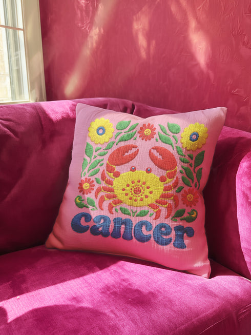 A square pink pillowcase features a vibrant, multi-colored design centered around a yellow and red crab representing the Cancer zodiac sign. The design is framed by symmetrical green leaves and yellow and red flowers. The word "cancer" is written in a bold, blue, rounded font at the bottom. The visual texture mimics intricate raised embroidery with visible stitch patterns, though the surface is a smooth 2D print on a linen-textured polyester fabric with a hidden zipper.