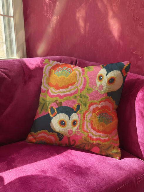 A square linen-feel throw pillow with a vibrant, maximalist print on a bright pink background. The pattern features two large, illustrated possums (or sugar gliders). The animals are nestled amongst large, 70s-style retro floral designs in shades of orange, marigold yellow, and turquoise, with avocado green leaves. The pillow has a textured, woven appearance, mimicking a high-end linen fabric. The double-sided print is colorful, eclectic, and groovy, with a distinct vintage 70s folk art feel.