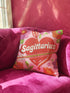 A square decorative throw pillow featuring a light pink background with a trompe-l'œil faux-embroidery print. A large red heart sits in the center, pierced by a green and white arrow. The word Sagittarius is printed across the heart in a puffy white retro font with a red drop shadow. The central design is surrounded by scattered vintage-style flowers in vibrant shades of purple, orange, and yellow with bright green leaves. The texture is a flat, high-fidelity printed illusion.