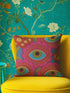 A square decorative throw pillow with a premium linen-feel texture. The design is a repeating pattern of stylized evil eyes on a dusty blue background. Each eye is rendered in turquoise, olive green, and black, and is framed by vibrant hot pink petals with radiating gold lines, resembling a flower or sunburst. The all-over print is colorful and has a visual texture that mimics embroidery. The pillow has a clean, knife-edge finish and a double-sided design, showcasing a bold, psychedelic pattern.