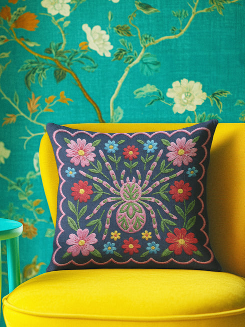 Square throw pillow cover featuring a symmetrical folk art spider illustration centered on a large hot pink flower. The spider has a striped abdomen in green, red, and yellow with pink legs. The background is a textured dark navy blue fabric with a visible faux linen weave. Surrounding the central spider are stylized botanical motifs including red daisies, blue forget-me-nots, and green leaves. The illustration is flat printed with a high-quality finish. This image shows the case only.