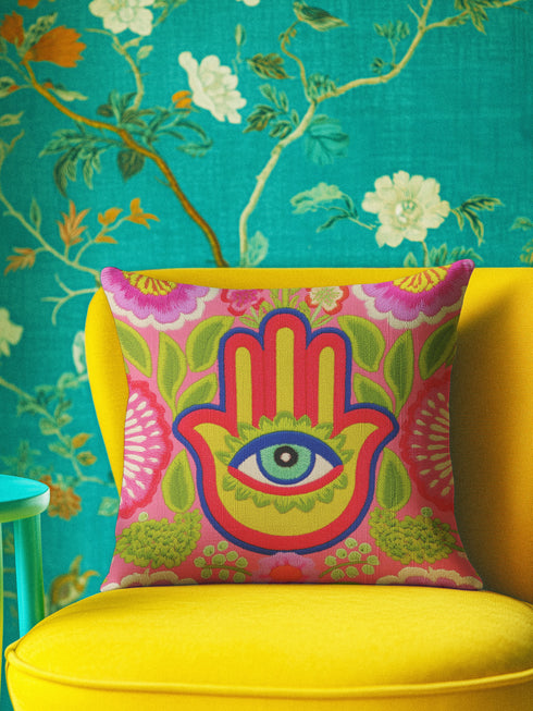 A square decorative throw pillow cover featuring a vibrant pink background with a bold, symmetrical design. In the center is a large, multi-colored Hamsa hand symbol outlined in red and yellow, containing a detailed evil eye with a green iris and black pupil. The hand is surrounded by stylized botanical elements, including pink flowers and green leaves. The design features a visual texture that mimics intricate thread embroidery but is a smooth, flat print on fabric.