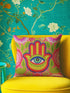 A square decorative throw pillow cover featuring a vibrant pink background with a bold, symmetrical design. In the center is a large, multi-colored Hamsa hand symbol outlined in red and yellow, containing a detailed evil eye with a green iris and black pupil. The hand is surrounded by stylized botanical elements, including pink flowers and green leaves. The design features a visual texture that mimics intricate thread embroidery but is a smooth, flat print on fabric.