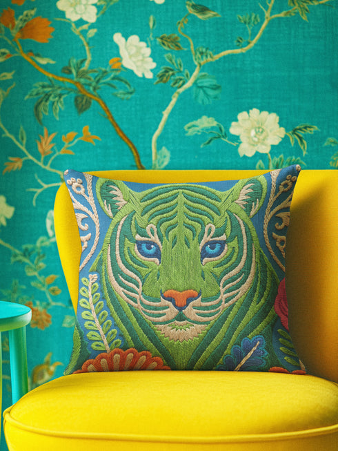 A square decorative throw pillow cover featuring a detailed illustration of a green tiger face in an Art Nouveau style. The tiger has piercing blue eyes and a bright orange nose, surrounded by intricate swirling botanical leaves in shades of teal and sage. The design mimics the look of embroidery or needlepoint but is a flat print on textured faux linen fabric. The background is a deep blue-green. The image is symmetrical and bold, fitting maximalist or junglecore aesthetics