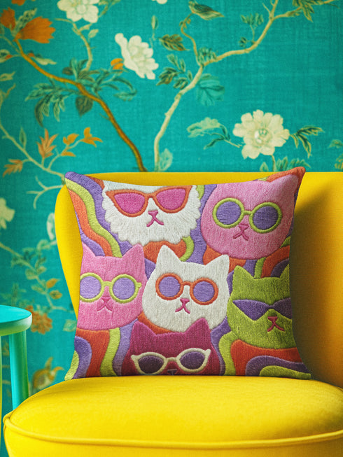 Square throw pillow cover packed with a dense repeating pattern of cat faces in hot pink, lime green, purple, and white, every cat wearing oversized retro sunglasses. Wavy psychedelic bands of orange, yellow, and green weave through the background. The flat 2D print mimics heavy textured crewel needlepoint on a smooth, linen-feel fabric. Reversible same design front and back with a hidden zipper. Funky maximalist cat decor and quirky apartment warming gift, available in 18 by 18 inch and 22 by 22 inch, cove