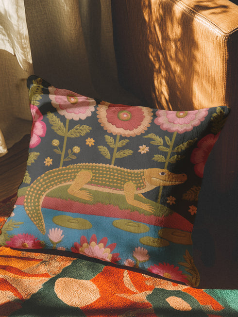 A vibrant square throw pillow cover featuring a green alligator illustration on a dark charcoal background. The design mimics the texture of vintage needlepoint embroidery on faux linen fabric. The alligator is accented with lime green scales and hot pink floral and lip motifs, creating a maximalist junglecore aesthetic. The pillow is shown on a furniture piece, highlighting the vivid cherry red and neon accents typical of dopamine decor and swampcore styles.