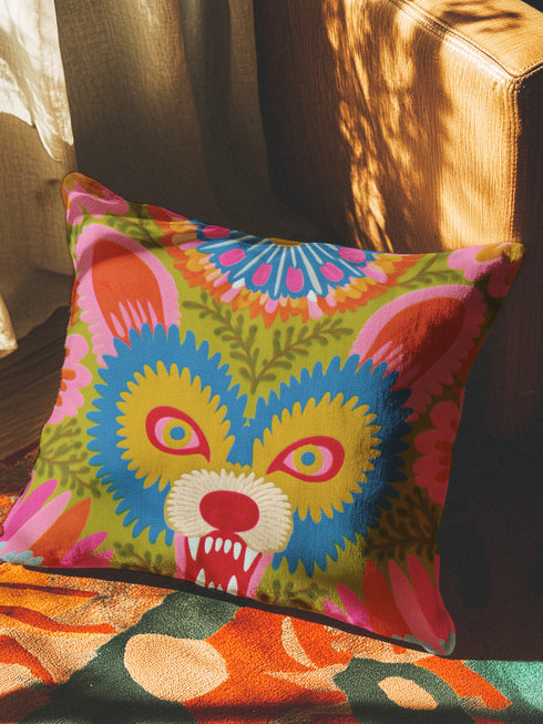 A square throw pillow cover featuring a vibrant, symmetrical illustration of an angry opossum face. The design mimics the texture of folk art embroidery but is a smooth flat print on faux linen fabric. The possum features teal blue fur, bright yellow eyes with pink pupils, and a red nose, bearing sharp white teeth in a hiss or scream. The background is a mossy green with pink and orange floral accents surrounding the animal. The style is maximalist, eclectic, colorful, and feral core aesthetic.