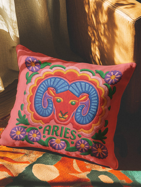 A square throw pillow featuring a vibrant, maximalist Aries zodiac design. The central motif is a red ram head with curled blue horns, surrounded by a scalloped border in yellow and pink. The design includes the word ARIES in green capital letters below the ram. Floral elements with purple flowers and green leaves frame the corners and bottom edge. The background is a textured coral-pink color. The artwork style is a 2D flat print designed to look like raised, textured embroidery.