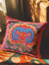 A square throw pillow featuring a vibrant, maximalist Aries zodiac design. The central motif is a red ram head with curled blue horns, surrounded by a scalloped border in yellow and pink. The design includes the word ARIES in green capital letters below the ram. Floral elements with purple flowers and green leaves frame the corners and bottom edge. The background is a textured coral-pink color. The artwork style is a 2D flat print designed to look like raised, textured embroidery.