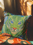 A square decorative throw pillow cover featuring a detailed illustration of a green tiger face in an Art Nouveau style. The tiger has piercing blue eyes and a bright orange nose, surrounded by intricate swirling botanical leaves in shades of teal and sage. The design mimics the look of embroidery or needlepoint but is a flat print on textured faux linen fabric. The background is a deep blue-green. The image is symmetrical and bold, fitting maximalist or junglecore aesthetics