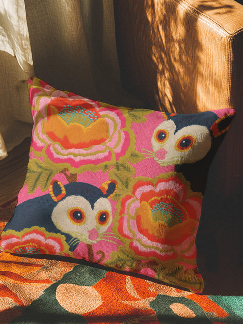 A square linen-feel throw pillow with a vibrant, maximalist print on a bright pink background. The pattern features two large, illustrated possums (or sugar gliders). The animals are nestled amongst large, 70s-style retro floral designs in shades of orange, marigold yellow, and turquoise, with avocado green leaves. The pillow has a textured, woven appearance, mimicking a high-end linen fabric. The double-sided print is colorful, eclectic, and groovy, with a distinct vintage 70s folk art feel.