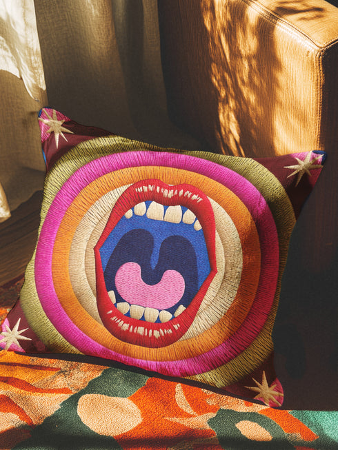 A square decorative pillowcase featuring a bold, centralized trompe-l'œil design printed to look like raised satin-stitch embroidery. The focal point is a large, open mouth with red lips, yellow teeth, and a pink tongue against a dark blue background. The mouth is surrounded by concentric radial rings in bright pink, orange, yellow, and olive green. The four corners are a dark burgundy brown, each accented with a yellow four-pointed starburst. The fabric has a woven linen-like texture.