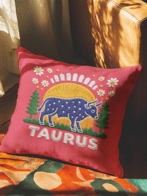 A square vibrant red throw pillow featuring a central illustration of the Taurus zodiac sign. The word TAURUS appears at the top in white bubbly letters with a faux-stitch texture. Below the text is an arch containing a purple bull standing in a green field dotted with white daisies and red flowers. Behind the bull are green pine trees and a large yellow sun against a blue sky. The design is framed by a pink wavy border, printed to look like raised embroidery on linen-textured polyester fabric.