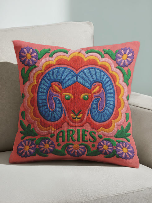 A square throw pillow featuring a vibrant, maximalist Aries zodiac design. The central motif is a red ram head with curled blue horns, surrounded by a scalloped border in yellow and pink. The design includes the word ARIES in green capital letters below the ram. Floral elements with purple flowers and green leaves frame the corners and bottom edge. The background is a textured coral-pink color. The artwork style is a 2D flat print designed to look like raised, textured embroidery.
