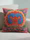 A square throw pillow featuring a vibrant, maximalist Aries zodiac design. The central motif is a red ram head with curled blue horns, surrounded by a scalloped border in yellow and pink. The design includes the word ARIES in green capital letters below the ram. Floral elements with purple flowers and green leaves frame the corners and bottom edge. The background is a textured coral-pink color. The artwork style is a 2D flat print designed to look like raised, textured embroidery.