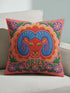 A square throw pillow featuring a vibrant, maximalist Aries zodiac design. The central motif is a red ram head with curled blue horns, surrounded by a scalloped border in yellow and pink. The design includes the word ARIES in green capital letters below the ram. Floral elements with purple flowers and green leaves frame the corners and bottom edge. The background is a textured coral-pink color. The artwork style is a 2D flat print designed to look like raised, textured embroidery.