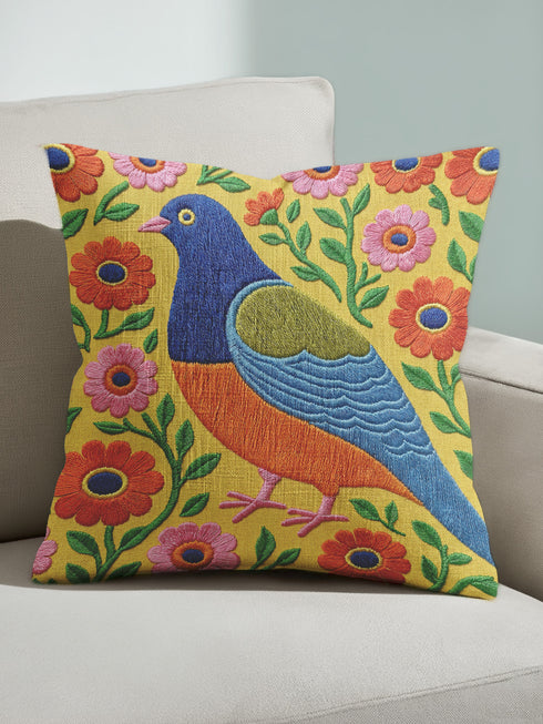 Square faux embroidery throw pillow cover with a large stylized folk art bird facing left. The bird has a cobalt blue head, orange belly, light blue tail, and pink legs, set on a bright mustard yellow background scattered with pink and red daisy flowers on trailing green vines. The smooth, linen-feel polyester uses a printed trompe loeil effect to mimic raised thread needlework. Bold maximalist folk art bird pillowcase and cottagecore cushion cover for eclectic, colorful, dopamine decor homes.