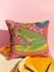 A square decorative throw pillow made from premium polyester with a visible linen-like woven texture. The background is a vibrant hot pink covered in a dense, retro-style floral pattern in orange, teal, and gold. The main design is a brightly colored, stylized illustration of a green red-eyed tree frog with orange feet and prominent red eyes. The whimsical frog is riding a vibrant yellow banana. The overall aesthetic is colorful, maximalist, and eclectic, with a faux-embroidered texture effect.