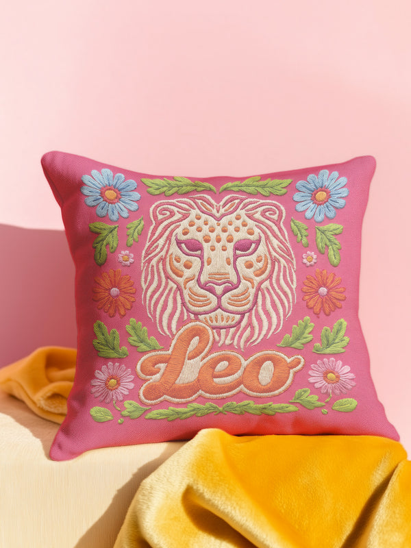 Maximalist throw pillow for the Leo zodiac sign featuring a centralized lion head medallion with an ornate mane mimicking embroidered textures. The background is a vibrant hot pink, accented by ochre, rust, and lime green floral motifs and blue flowers in each corner. The pillow is square with a hidden zipper and a polyester insert. This high-contrast design is a perfect fit for dopamine decor, whimsigoth apartments, or any eclectic home looking for a bold astrology-inspired statement piece.