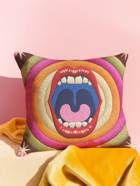 A square decorative pillowcase featuring a bold, centralized trompe-l'œil design printed to look like raised satin-stitch embroidery. The focal point is a large, open mouth with red lips, yellow teeth, and a pink tongue against a dark blue background. The mouth is surrounded by concentric radial rings in bright pink, orange, yellow, and olive green. The four corners are a dark burgundy brown, each accented with a yellow four-pointed starburst. The fabric has a woven linen-like texture.