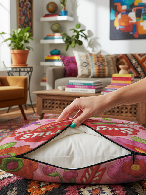 A square decorative throw pillow featuring a light pink background with a trompe-l'œil faux-embroidery print. A large red heart sits in the center, pierced by a green and white arrow. The word Sagittarius is printed across the heart in a puffy white retro font with a red drop shadow. The central design is surrounded by scattered vintage-style flowers in vibrant shades of purple, orange, and yellow with bright green leaves. The texture is a flat, high-fidelity printed illusion.