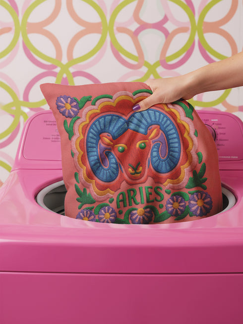 A square throw pillow featuring a vibrant, maximalist Aries zodiac design. The central motif is a red ram head with curled blue horns, surrounded by a scalloped border in yellow and pink. The design includes the word ARIES in green capital letters below the ram. Floral elements with purple flowers and green leaves frame the corners and bottom edge. The background is a textured coral-pink color. The artwork style is a 2D flat print designed to look like raised, textured embroidery.