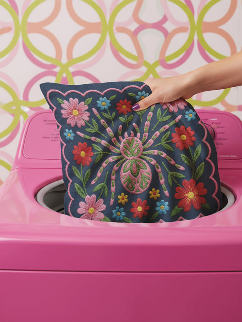 Square throw pillow cover featuring a symmetrical folk art spider illustration centered on a large hot pink flower. The spider has a striped abdomen in green, red, and yellow with pink legs. The background is a textured dark navy blue fabric with a visible faux linen weave. Surrounding the central spider are stylized botanical motifs including red daisies, blue forget-me-nots, and green leaves. The illustration is flat printed with a high-quality finish. This image shows the case only.