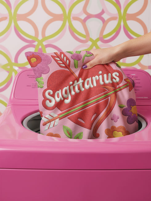 A square decorative throw pillow featuring a light pink background with a trompe-l'œil faux-embroidery print. A large red heart sits in the center, pierced by a green and white arrow. The word Sagittarius is printed across the heart in a puffy white retro font with a red drop shadow. The central design is surrounded by scattered vintage-style flowers in vibrant shades of purple, orange, and yellow with bright green leaves. The texture is a flat, high-fidelity printed illusion.