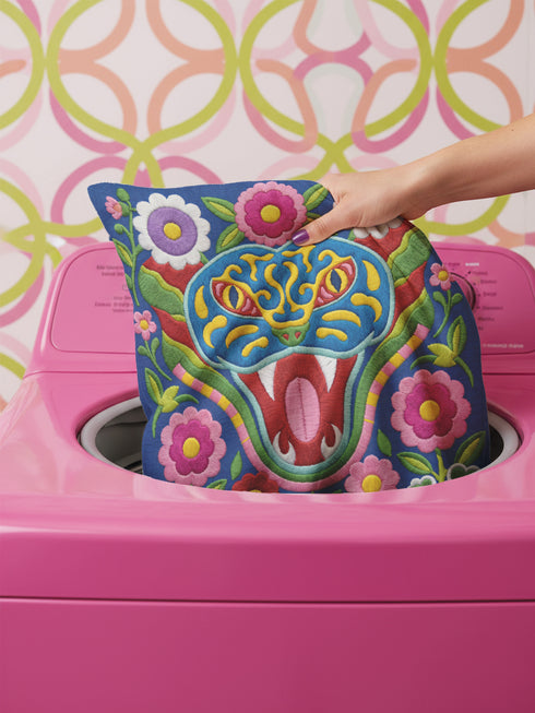 Square throw pillow cover with a faux embroidery 2D print of a stylized folk-art serpent with a blue head, yellow swirl markings, red eyes, white fangs, and an open red mouth. It is surrounded by pink, purple, and white flowers with yellow centers and green leaves on a deep blue background. The print mimics raised stitching while staying flat and smooth, and shows on both sides. Cover only, available in 18 by 18 inch and 22 by 22 inch sizes.