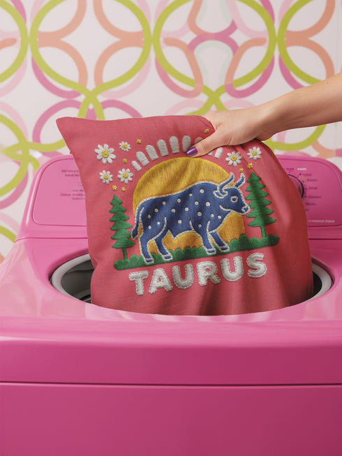 A square vibrant red throw pillow featuring a central illustration of the Taurus zodiac sign. The word TAURUS appears at the top in white bubbly letters with a faux-stitch texture. Below the text is an arch containing a purple bull standing in a green field dotted with white daisies and red flowers. Behind the bull are green pine trees and a large yellow sun against a blue sky. The design is framed by a pink wavy border, printed to look like raised embroidery on linen-textured polyester fabric.