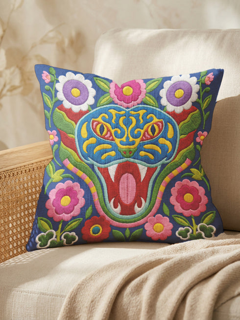 Square throw pillow cover with a faux embroidery 2D print of a stylized folk-art serpent with a blue head, yellow swirl markings, red eyes, white fangs, and an open red mouth. It is surrounded by pink, purple, and white flowers with yellow centers and green leaves on a deep blue background. The print mimics raised stitching while staying flat and smooth, and shows on both sides. Cover only, available in 18 by 18 inch and 22 by 22 inch sizes.