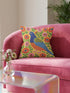 Square faux embroidery throw pillow cover with a large stylized folk art bird facing left. The bird has a cobalt blue head, orange belly, light blue tail, and pink legs, set on a bright mustard yellow background scattered with pink and red daisy flowers on trailing green vines. The smooth, linen-feel polyester uses a printed trompe loeil effect to mimic raised thread needlework. Bold maximalist folk art bird pillowcase and cottagecore cushion cover for eclectic, colorful, dopamine decor homes.