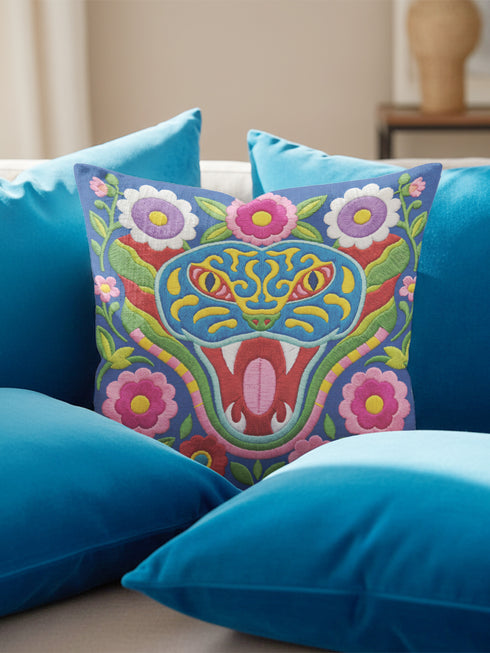 Square throw pillow cover with a faux embroidery 2D print of a stylized folk-art serpent with a blue head, yellow swirl markings, red eyes, white fangs, and an open red mouth. It is surrounded by pink, purple, and white flowers with yellow centers and green leaves on a deep blue background. The print mimics raised stitching while staying flat and smooth, and shows on both sides. Cover only, available in 18 by 18 inch and 22 by 22 inch sizes.