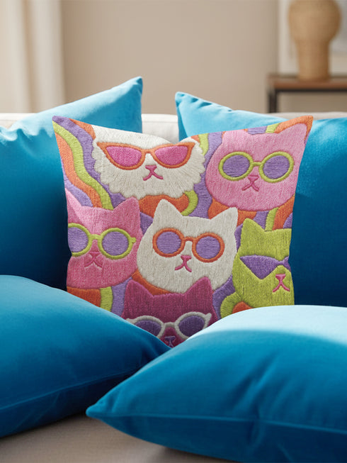Square throw pillow cover packed with a dense repeating pattern of cat faces in hot pink, lime green, purple, and white, every cat wearing oversized retro sunglasses. Wavy psychedelic bands of orange, yellow, and green weave through the background. The flat 2D print mimics heavy textured crewel needlepoint on a smooth, linen-feel fabric. Reversible same design front and back with a hidden zipper. Funky maximalist cat decor and quirky apartment warming gift, available in 18 by 18 inch and 22 by 22 inch, cove
