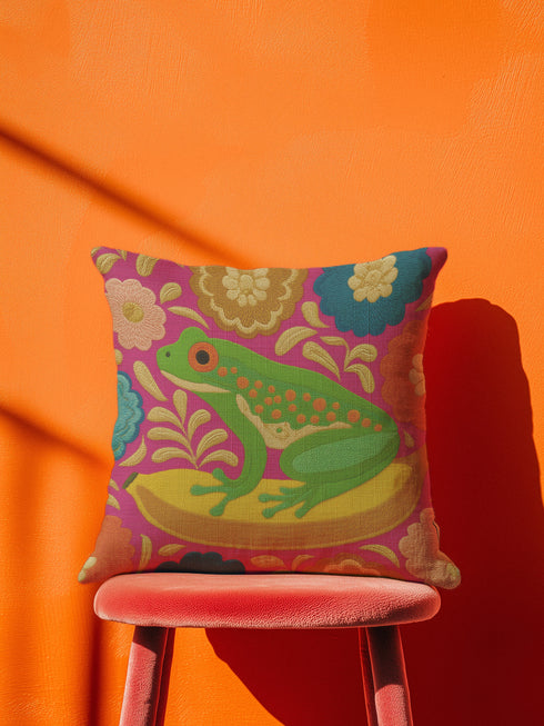 A square decorative throw pillow made from premium polyester with a visible linen-like woven texture. The background is a vibrant hot pink covered in a dense, retro-style floral pattern in orange, teal, and gold. The main design is a brightly colored, stylized illustration of a green red-eyed tree frog with orange feet and prominent red eyes. The whimsical frog is riding a vibrant yellow banana. The overall aesthetic is colorful, maximalist, and eclectic, with a faux-embroidered texture effect.