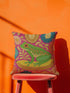 A square decorative throw pillow made from premium polyester with a visible linen-like woven texture. The background is a vibrant hot pink covered in a dense, retro-style floral pattern in orange, teal, and gold. The main design is a brightly colored, stylized illustration of a green red-eyed tree frog with orange feet and prominent red eyes. The whimsical frog is riding a vibrant yellow banana. The overall aesthetic is colorful, maximalist, and eclectic, with a faux-embroidered texture effect.