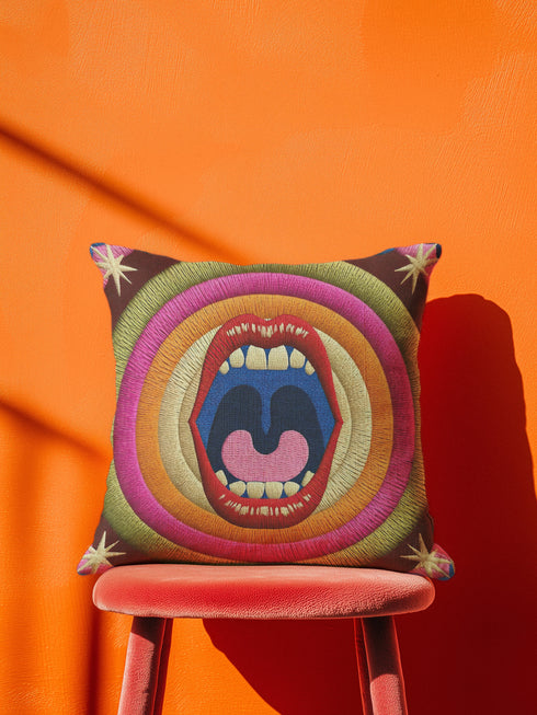 A square decorative pillowcase featuring a bold, centralized trompe-l'œil design printed to look like raised satin-stitch embroidery. The focal point is a large, open mouth with red lips, yellow teeth, and a pink tongue against a dark blue background. The mouth is surrounded by concentric radial rings in bright pink, orange, yellow, and olive green. The four corners are a dark burgundy brown, each accented with a yellow four-pointed starburst. The fabric has a woven linen-like texture.