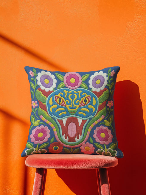 Square throw pillow cover with a faux embroidery 2D print of a stylized folk-art serpent with a blue head, yellow swirl markings, red eyes, white fangs, and an open red mouth. It is surrounded by pink, purple, and white flowers with yellow centers and green leaves on a deep blue background. The print mimics raised stitching while staying flat and smooth, and shows on both sides. Cover only, available in 18 by 18 inch and 22 by 22 inch sizes.