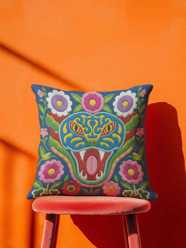 Square throw pillow cover with a faux embroidery 2D print of a stylized folk-art serpent with a blue head, yellow swirl markings, red eyes, white fangs, and an open red mouth. It is surrounded by pink, purple, and white flowers with yellow centers and green leaves on a deep blue background. The print mimics raised stitching while staying flat and smooth, and shows on both sides. Cover only, available in 18 by 18 inch and 22 by 22 inch sizes.