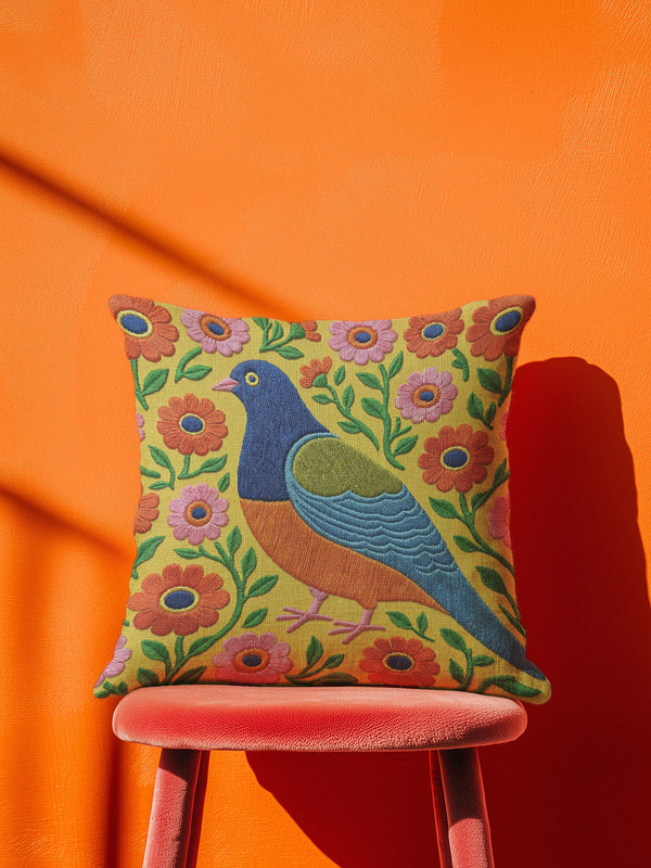 Square faux embroidery throw pillow cover with a large stylized folk art bird facing left. The bird has a cobalt blue head, orange belly, light blue tail, and pink legs, set on a bright mustard yellow background scattered with pink and red daisy flowers on trailing green vines. The smooth, linen-feel polyester uses a printed trompe loeil effect to mimic raised thread needlework. Bold maximalist folk art bird pillowcase and cottagecore cushion cover for eclectic, colorful, dopamine decor homes.