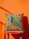 Square faux embroidery throw pillow cover with a large stylized folk art bird facing left. The bird has a cobalt blue head, orange belly, light blue tail, and pink legs, set on a bright mustard yellow background scattered with pink and red daisy flowers on trailing green vines. The smooth, linen-feel polyester uses a printed trompe loeil effect to mimic raised thread needlework. Bold maximalist folk art bird pillowcase and cottagecore cushion cover for eclectic, colorful, dopamine decor homes.